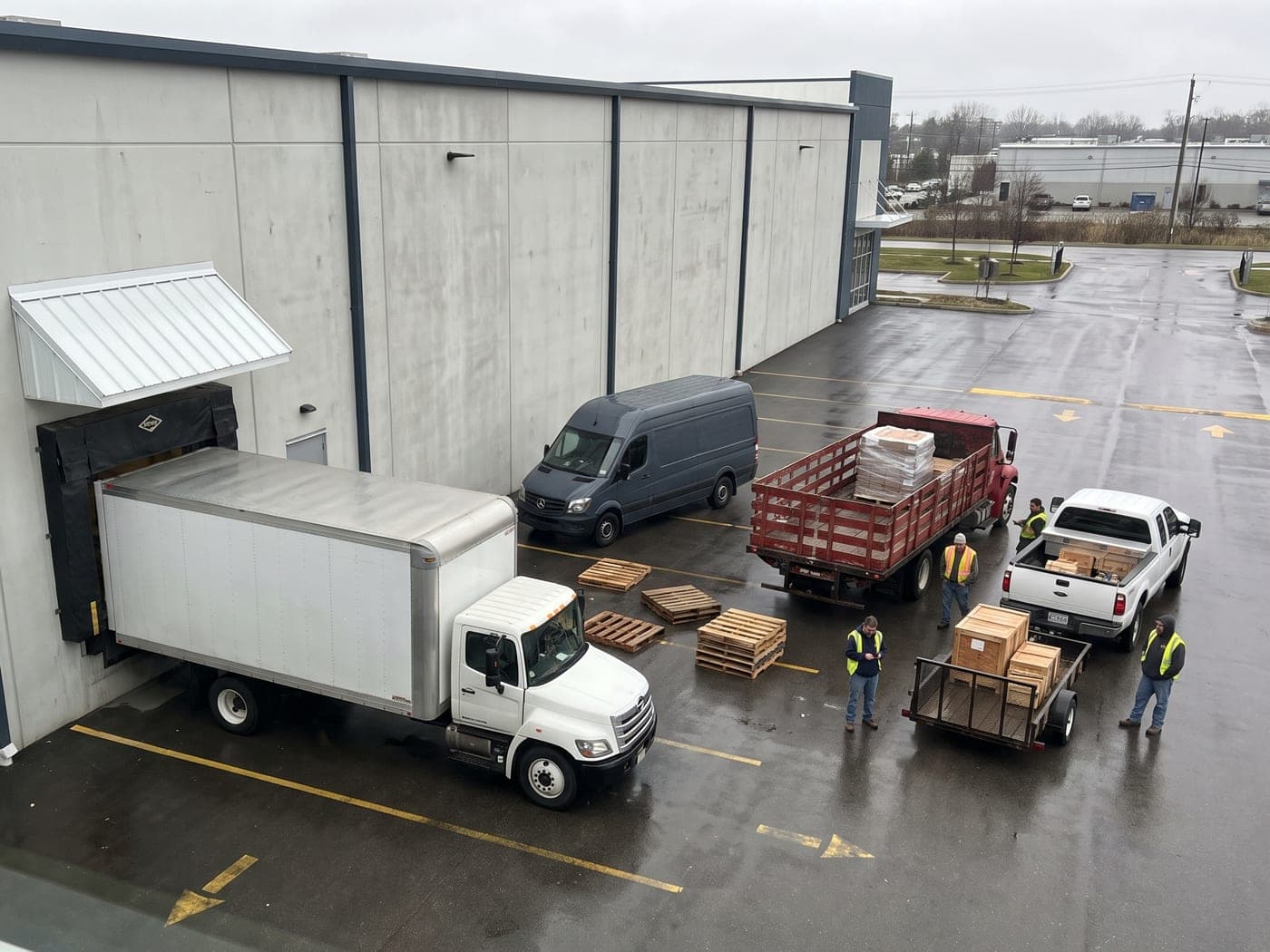 Looking down on a warehouse yard: a box truck at the only dock, with a cargo van, a flatbed and a pickup towing a trailer waiting out on the asphalt