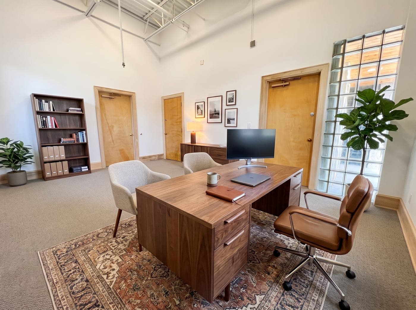 A well furnished private office with a walnut desk, leather chair and rug, lit by a tall glass block window wall