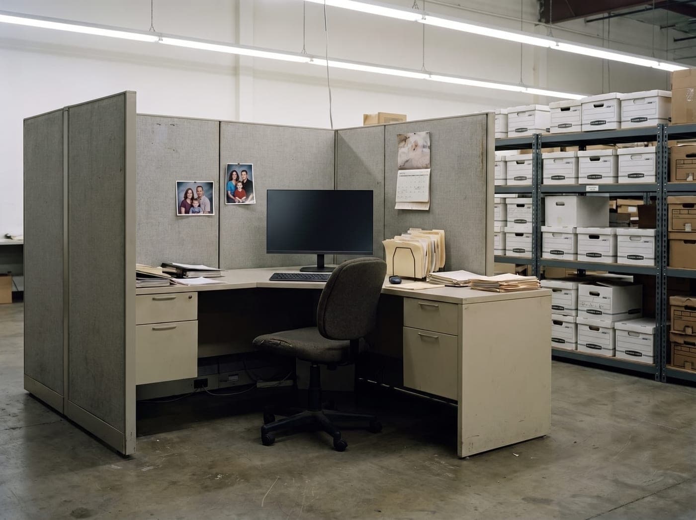 An old fabric cubicle in a windowless warehouse, lit by fluorescents, with racks of file boxes alongside