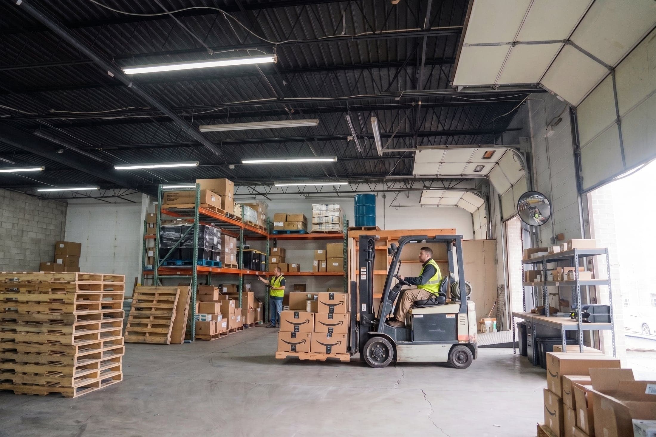Inside a leased small-bay warehouse: pallet racking, stacked cartons, a forklift moving an order, and the overhead door open to daylight